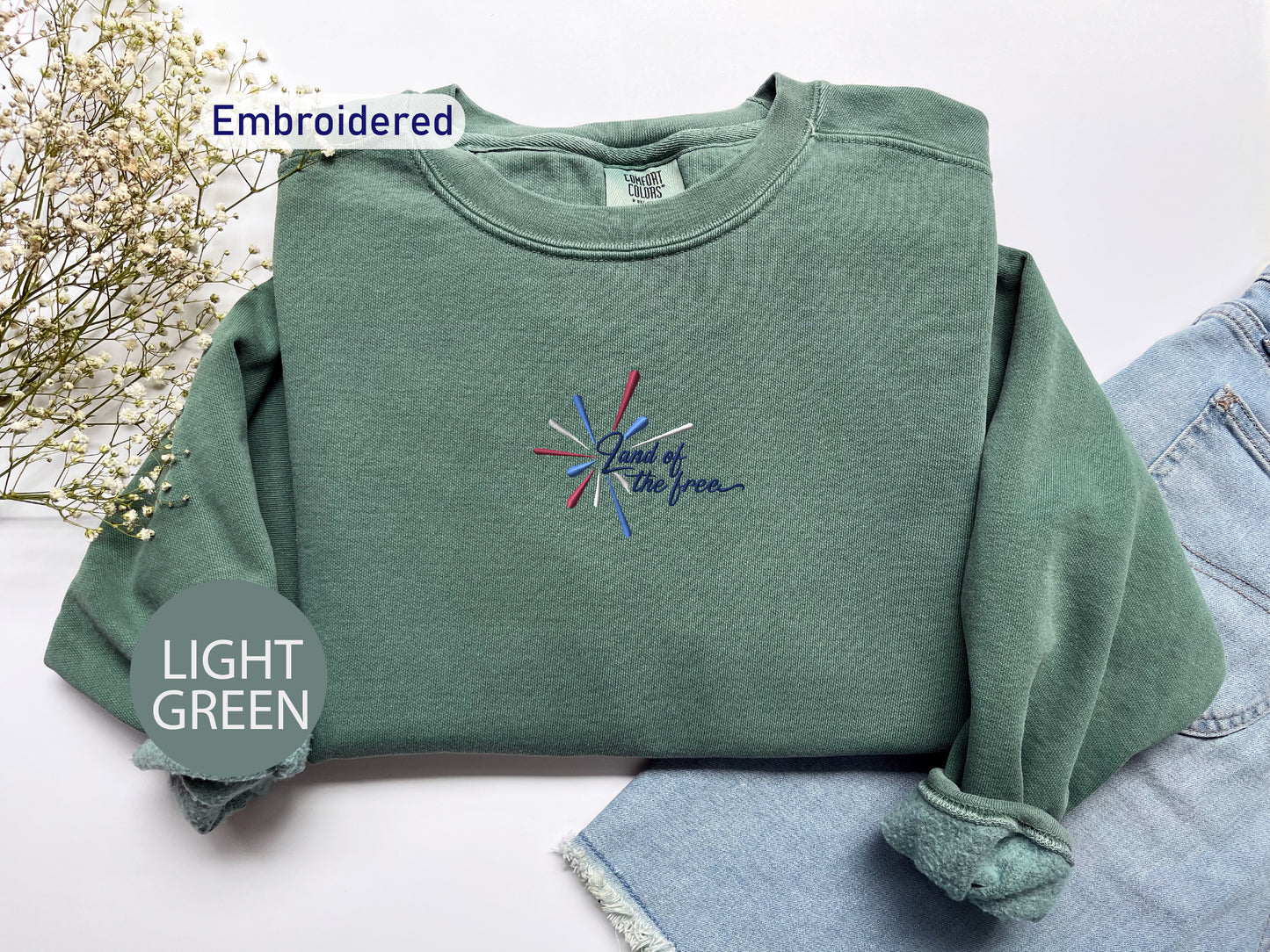 a green sweatshirt with a colorful embroidered design on it, placed on a white surface alongside a pair of denim jeans.