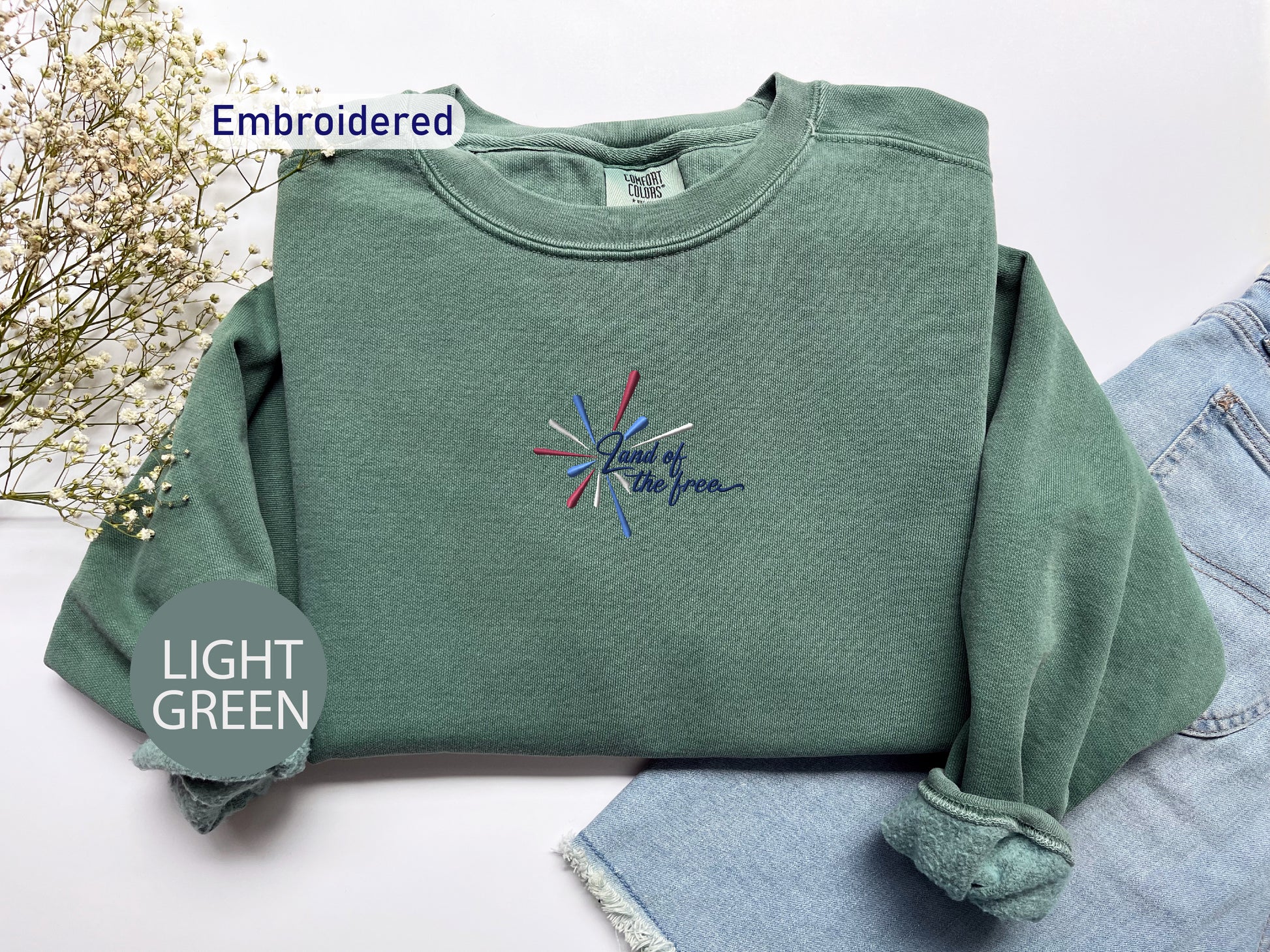 a green sweatshirt with a colorful embroidered design on it, placed on a white surface alongside a pair of denim jeans.