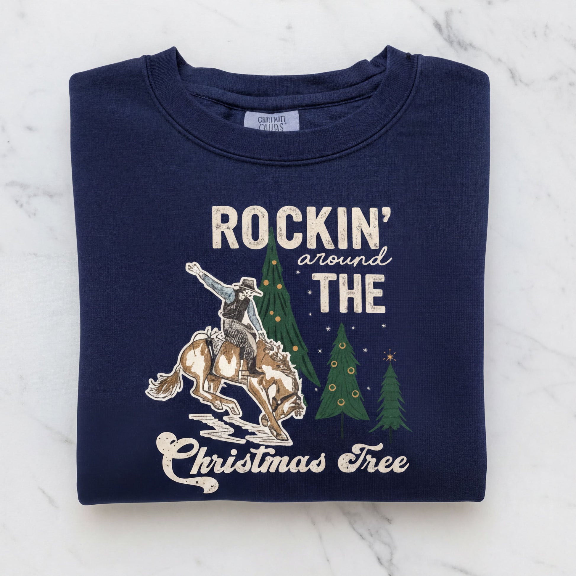 A navy blue sweatshirt with a graphic design depicting a cowboy riding a bucking bronco, with Christmas trees in the background. The text on the sweatshirt reads "ROCKIN' around THE CHRISTMAS Tree".