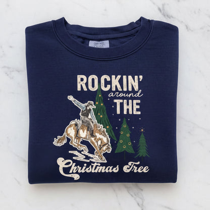 A navy blue sweatshirt with a graphic design depicting a cowboy riding a bucking bronco, with Christmas trees in the background. The text on the sweatshirt reads "ROCKIN' around THE CHRISTMAS Tree".