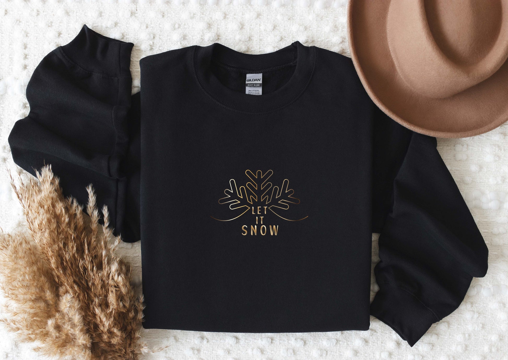 A black long-sleeved shirt with a gold snowflake design and the text "LET IT SNOW" printed on it, along with a beige hat and some dried plants.