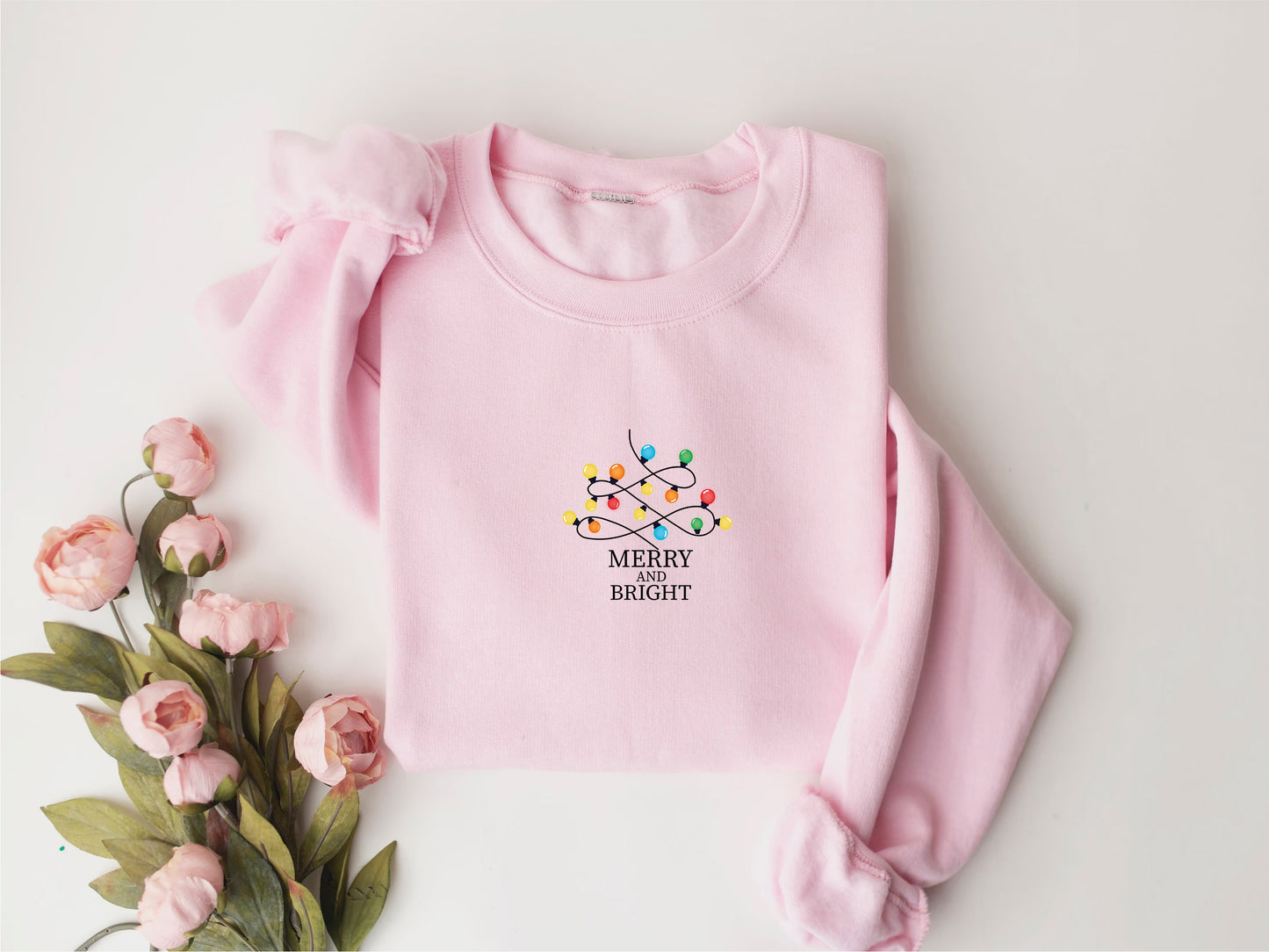 A pink sweater with a Christmas-themed design and the text "MERRY BRIOT" is displayed on a white background, accompanied by a bouquet of pink flowers.