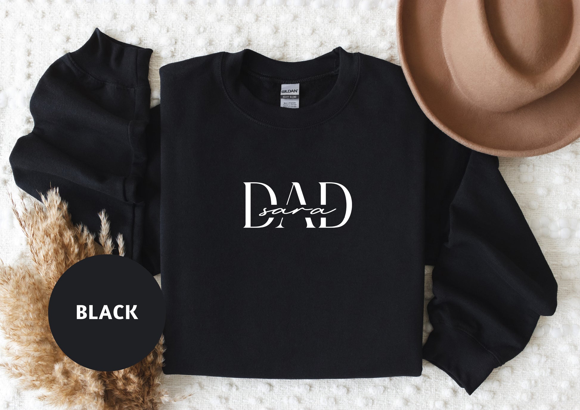 A black sweatshirt with the text "DAD" printed on it, along with a brown hat and some dried plants, all laid out on a white surface.