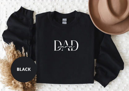 A black sweatshirt with the text "DAD" printed on it, along with a brown hat and some dried plants, all laid out on a white surface.