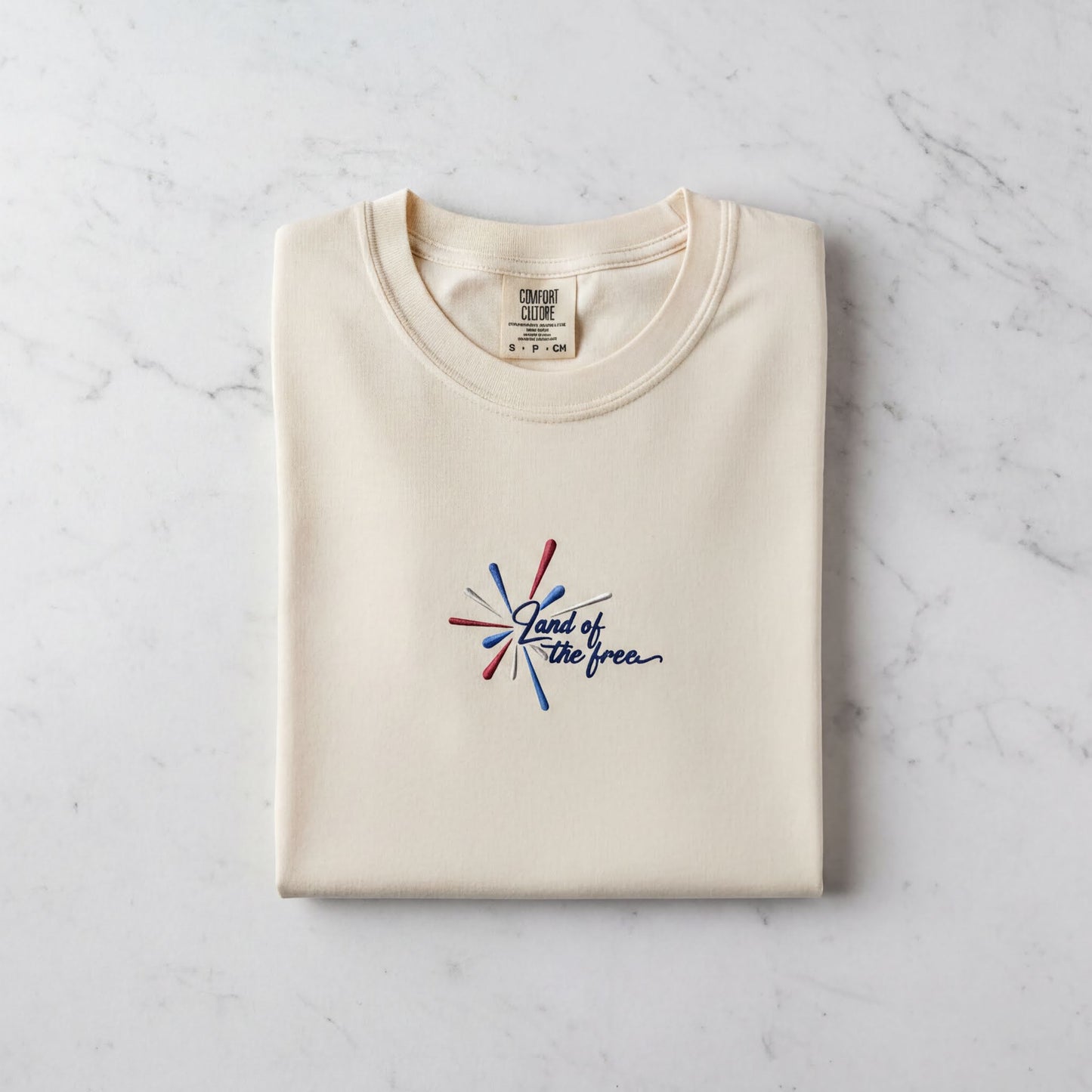 A cream-colored t-shirt with a graphic design and text printed on it, laid out on a marble surface.