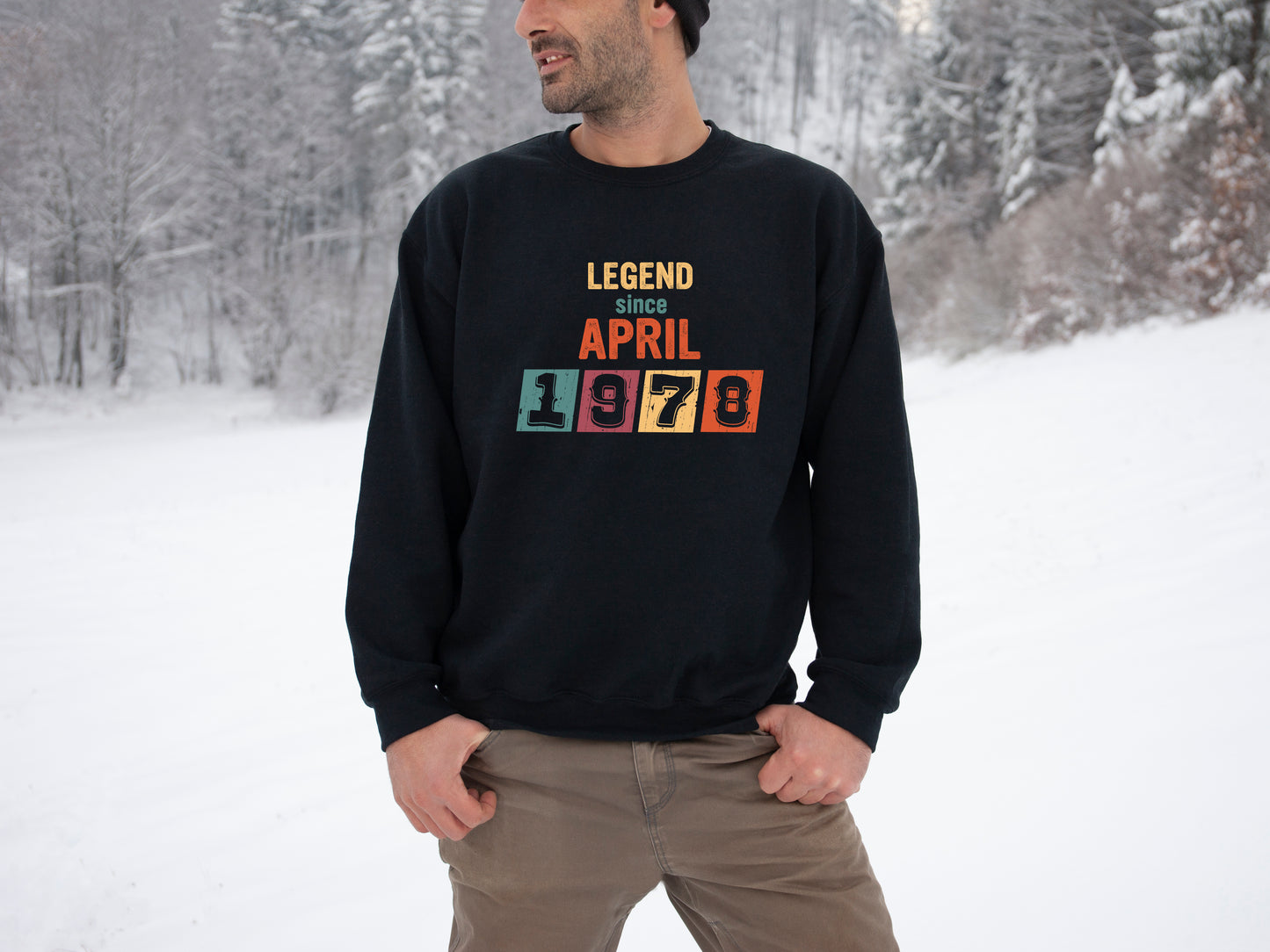 A man wearing a black sweatshirt with the text "LEGEND since APRIL 1978" printed on it, standing in a snowy forest.