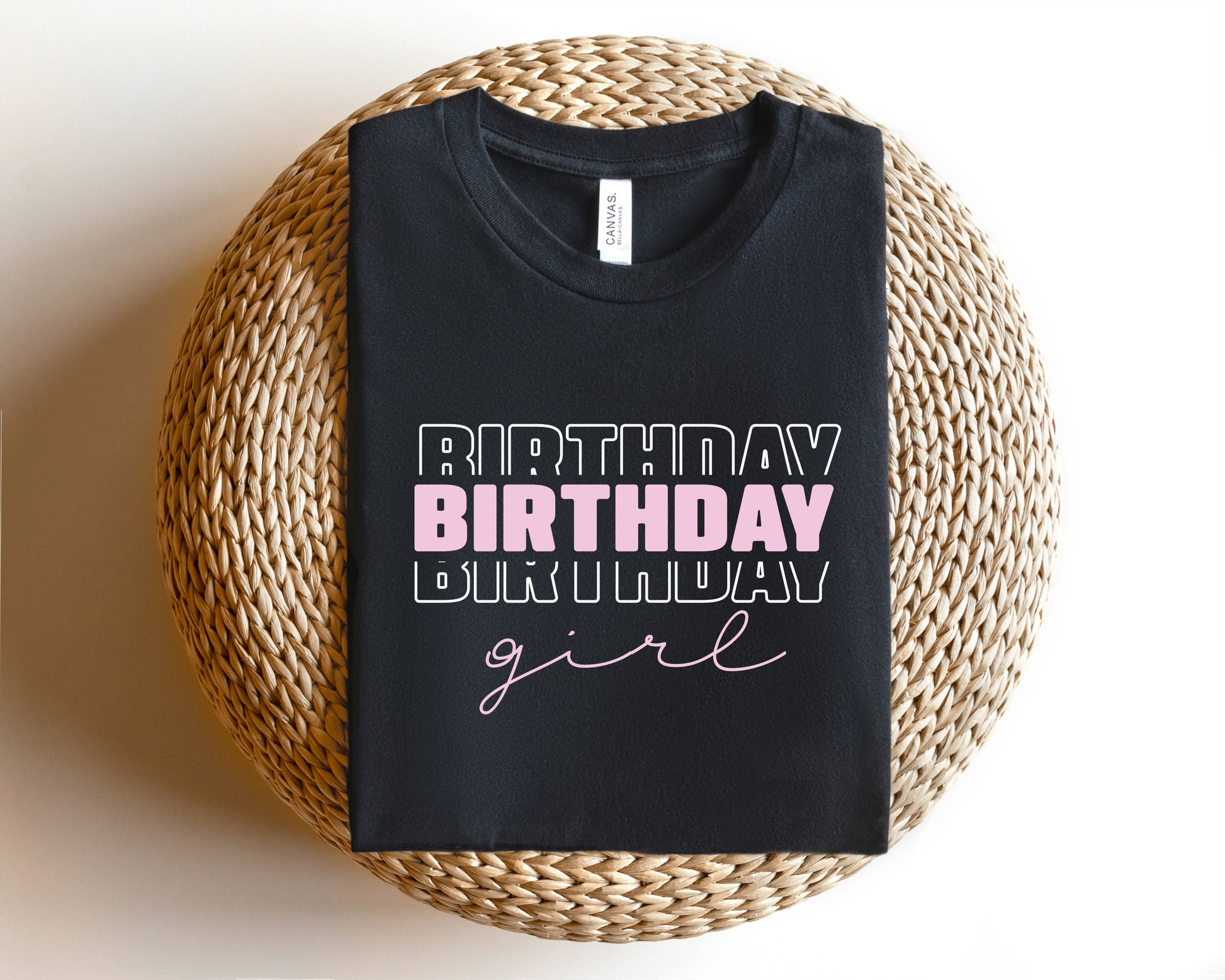 A black t-shirt with the text "BIRTHDAY GIRL" printed on it, placed on a woven basket or round pillow.