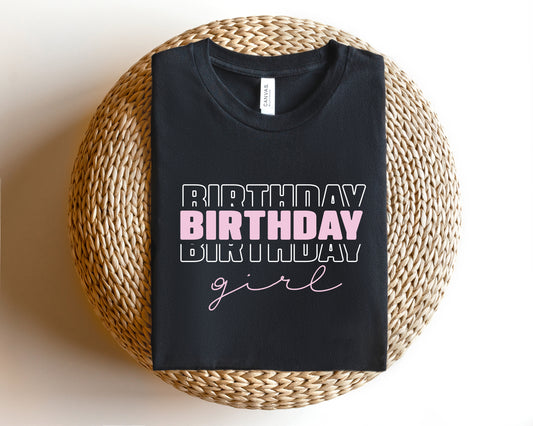 A black t-shirt with the text "BIRTHDAY GIRL" printed on it, placed on a woven basket or round pillow.