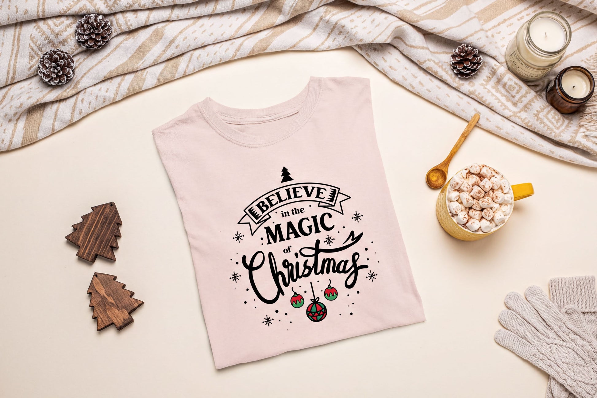 A pink t-shirt with a Christmas-themed design, including a tree, snowflakes, and ornaments, along with a mug of hot chocolate and pine cones, all arranged on a beige background.