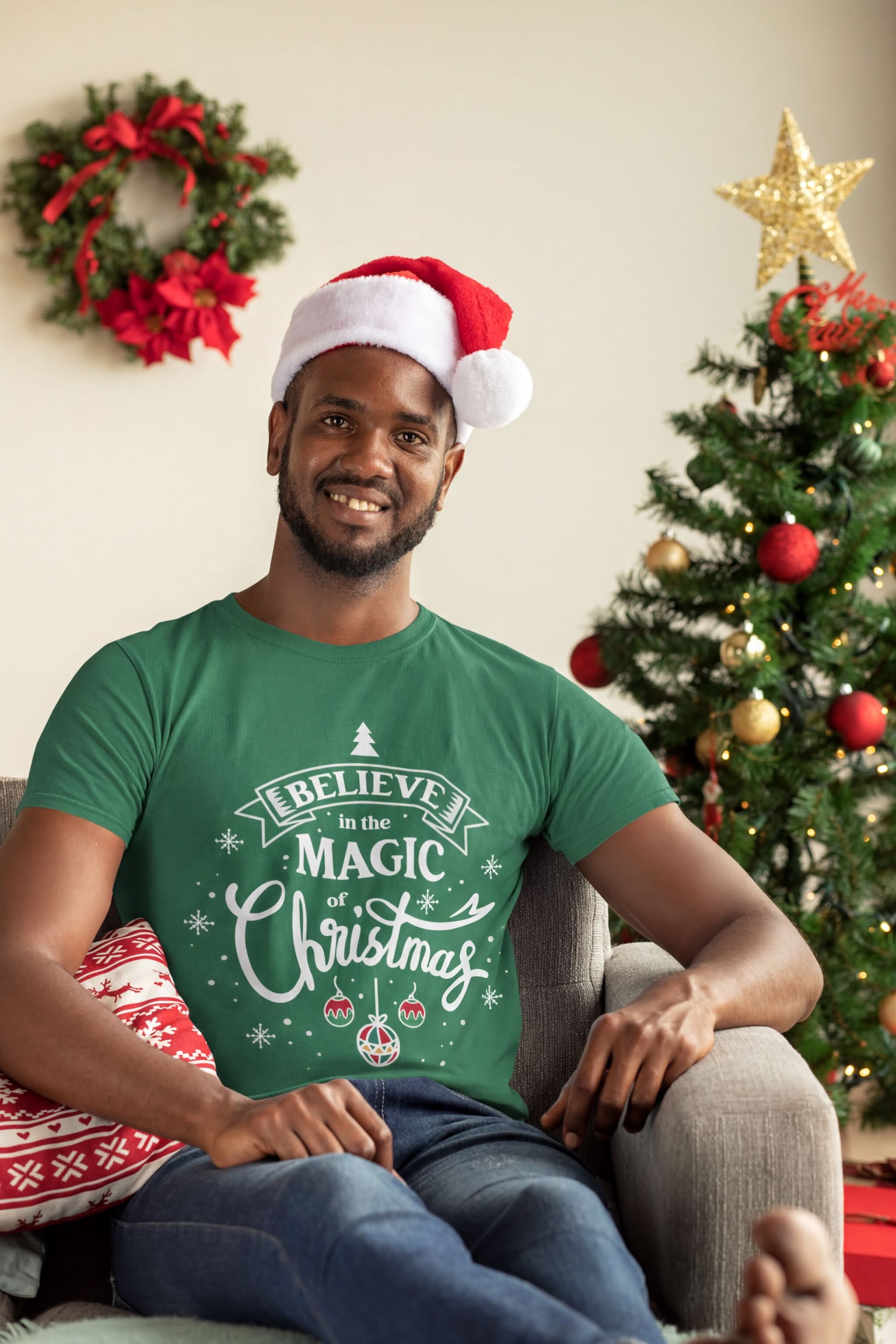 A smiling man wearing a Santa hat and a green t-shirt with a Christmas-themed design is sitting in a chair in front of a Christmas tree.
