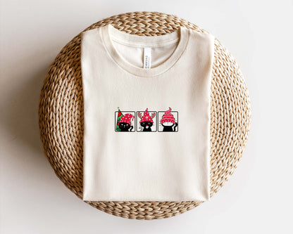 A white t-shirt with a graphic design of three cats wearing red hats, arranged in a row.