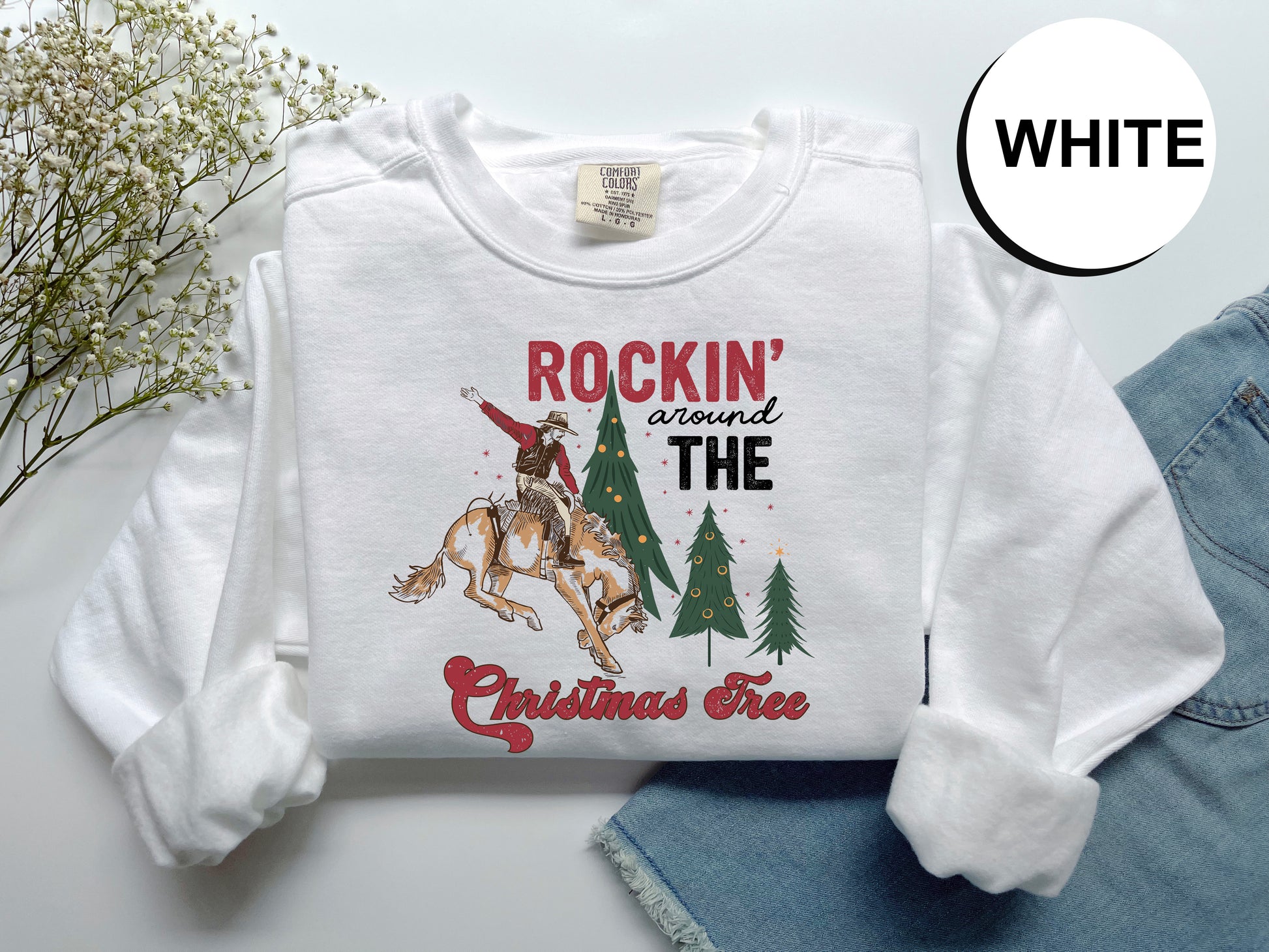 A white long-sleeved t-shirt with a graphic design featuring a cowboy riding a bucking bronco, surrounded by Christmas trees, and the text "Rockin' around the Christmas Tree".
