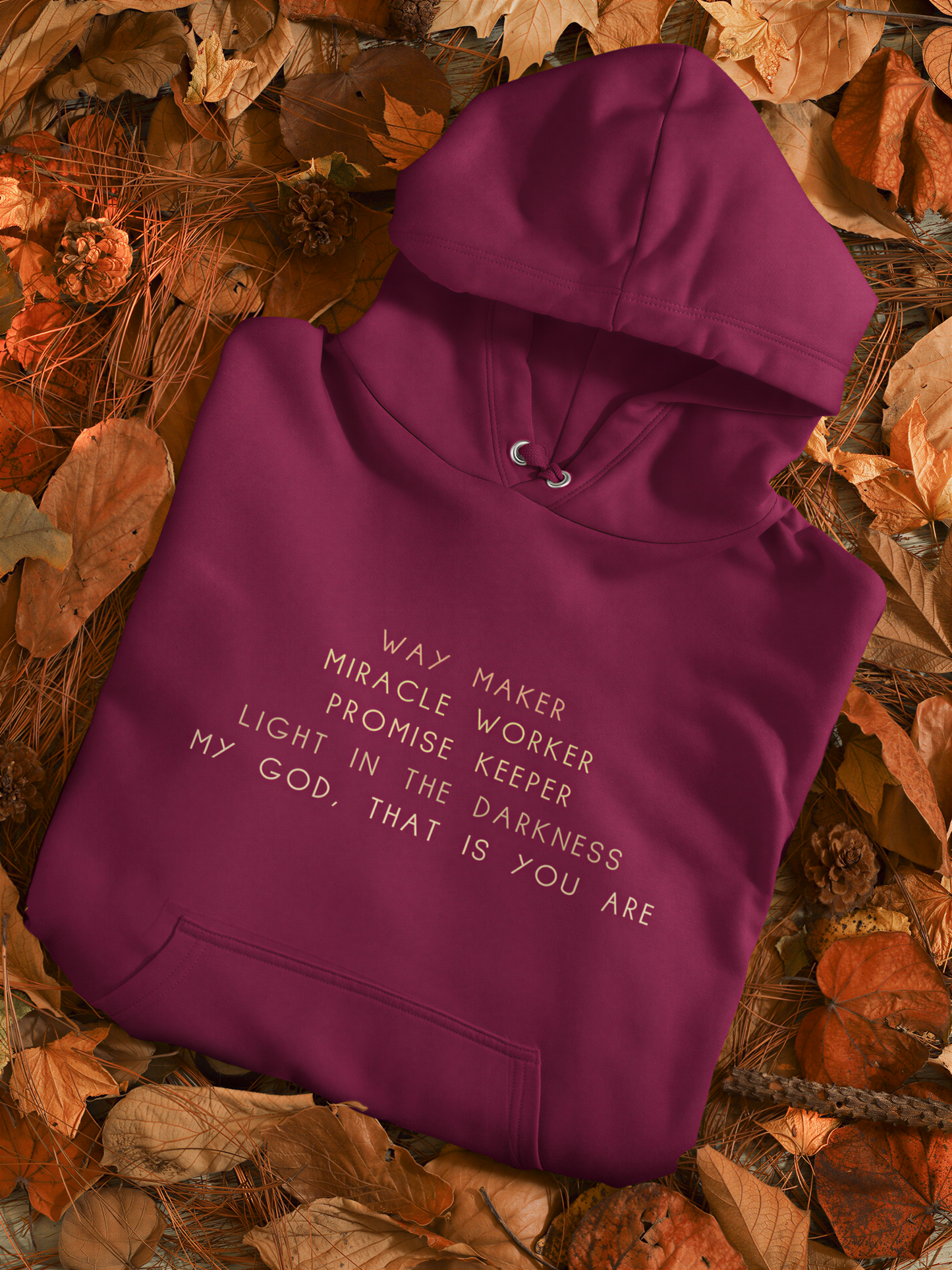 A maroon hoodie with white text is laid out on a bed of autumn leaves.