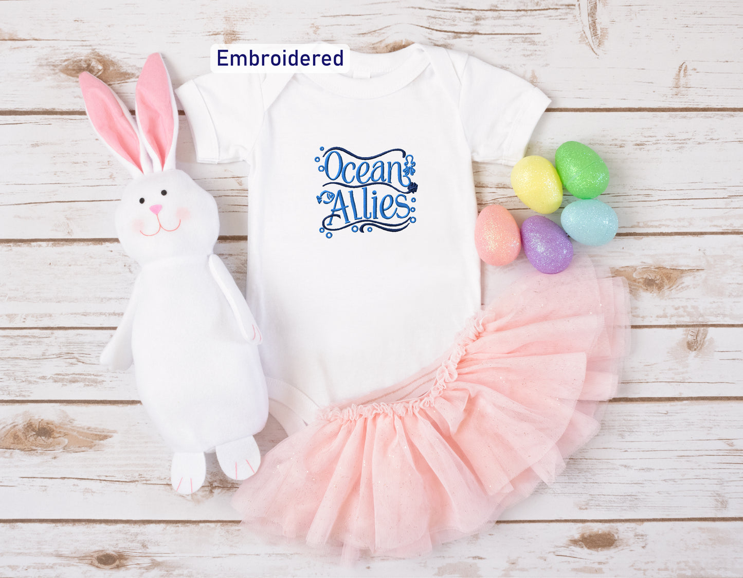 a white onesie with a blue embroidered design, a pink tulle skirt, a stuffed bunny, and colorful Easter eggs arranged on a white wooden surface.