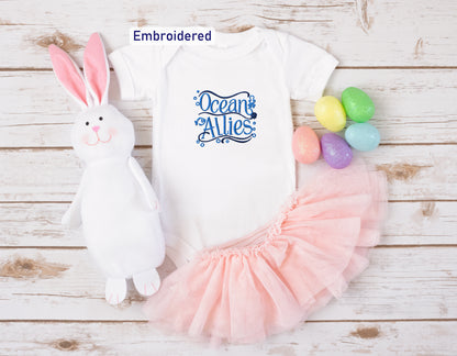 a white onesie with a blue embroidered design, a pink tulle skirt, a stuffed bunny, and colorful Easter eggs arranged on a white wooden surface.