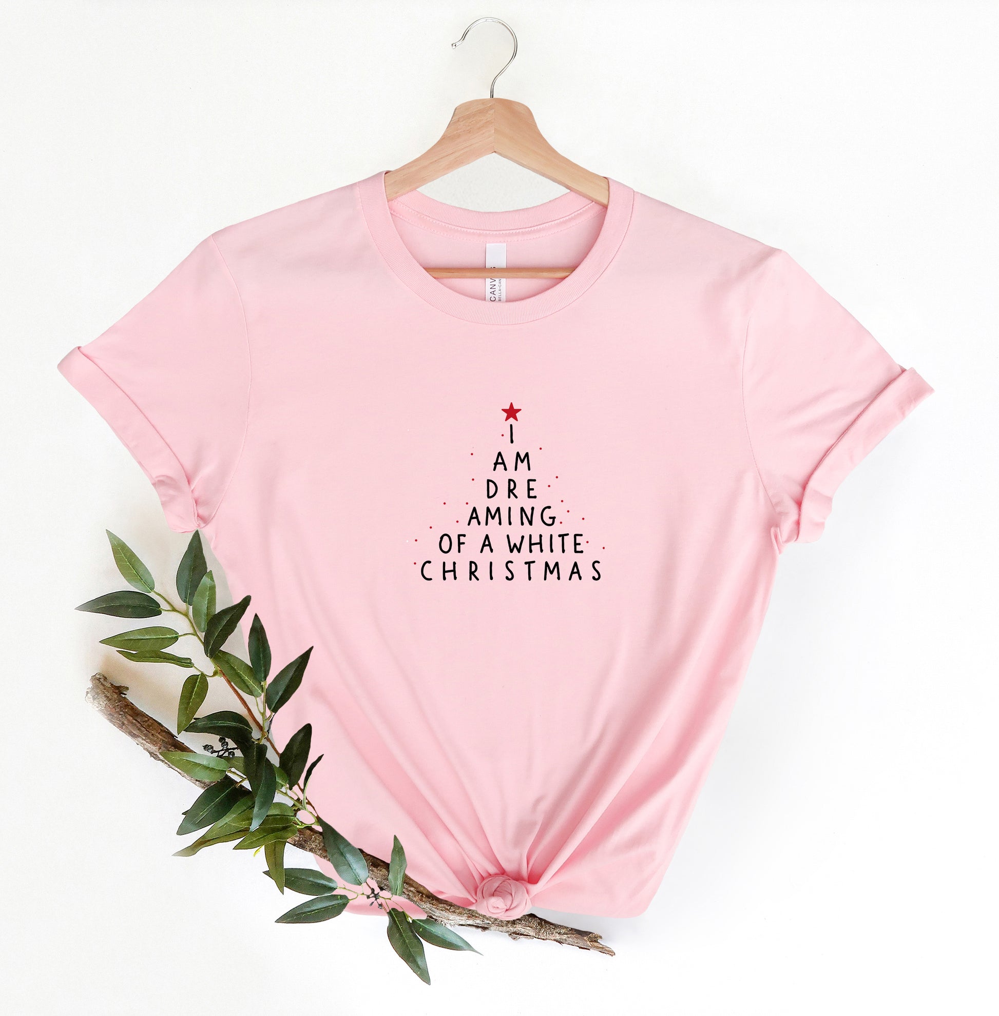 A pink t-shirt with a Christmas tree design and text printed on it, hanging on a wooden hanger.