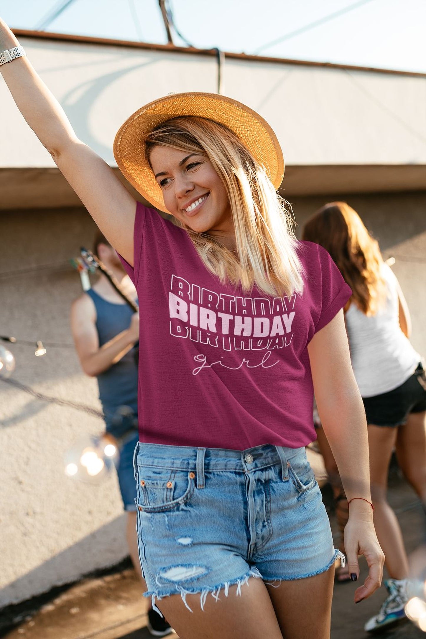 A smiling woman wearing a pink t-shirt with the text "BIRTHDAY GIRL" and denim shorts, along with a straw hat, is standing outdoors in front of a building.