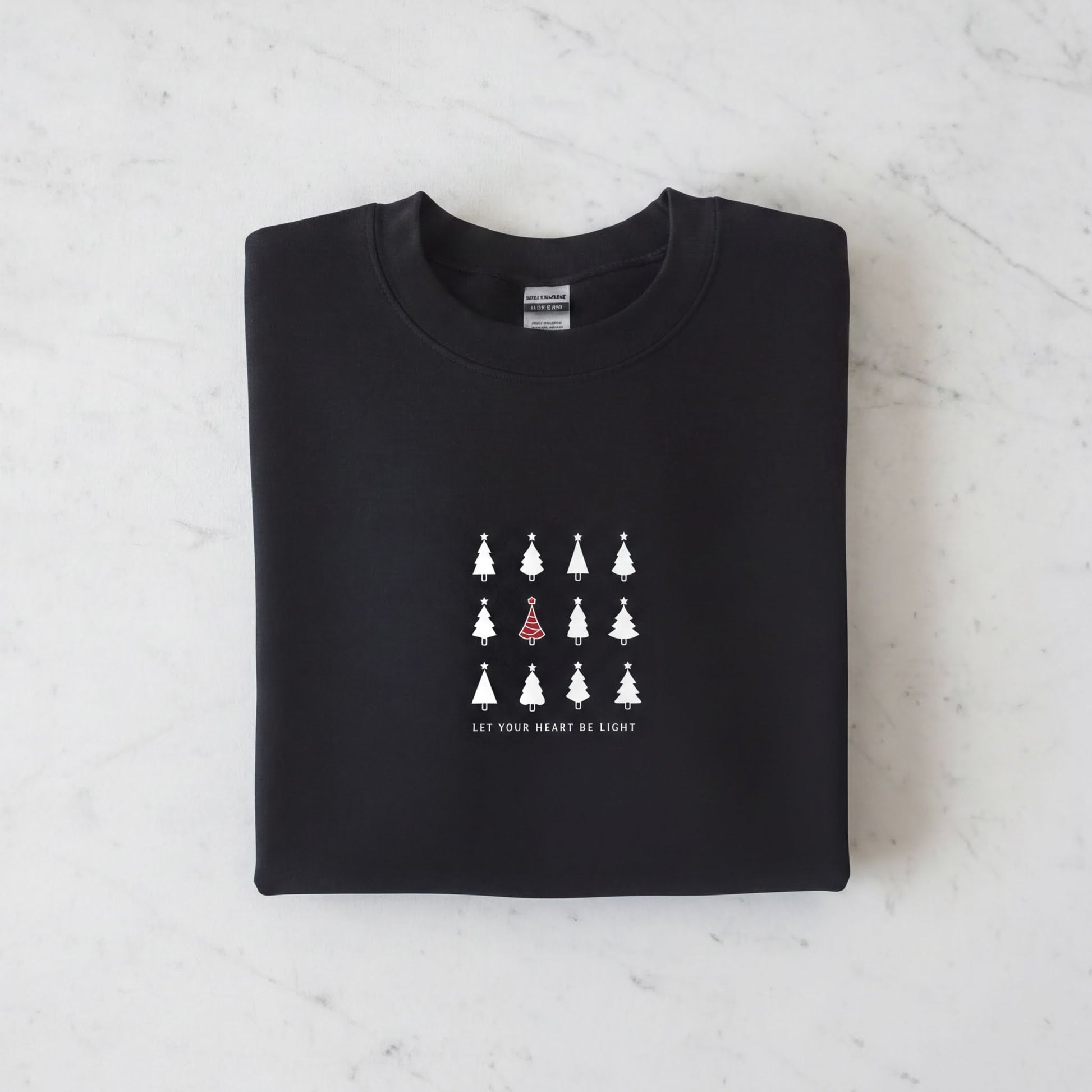 A black sweatshirt with a white graphic design on the front. The design consists of a grid of white Christmas trees, with one tree standing out in red. The phrase "Let your heart be light" is printed below the graphic.