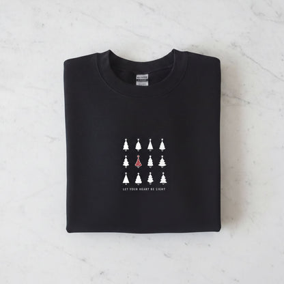 A black sweatshirt with a white graphic design on the front. The design consists of a grid of white Christmas trees, with one tree standing out in red. The phrase "Let your heart be light" is printed below the graphic.