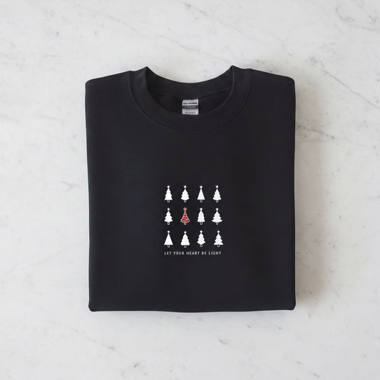 A black sweatshirt with a white graphic design on the front. The design consists of a grid of white Christmas trees, with one tree standing out in red. The phrase "Let your heart be light" is printed below the graphic.