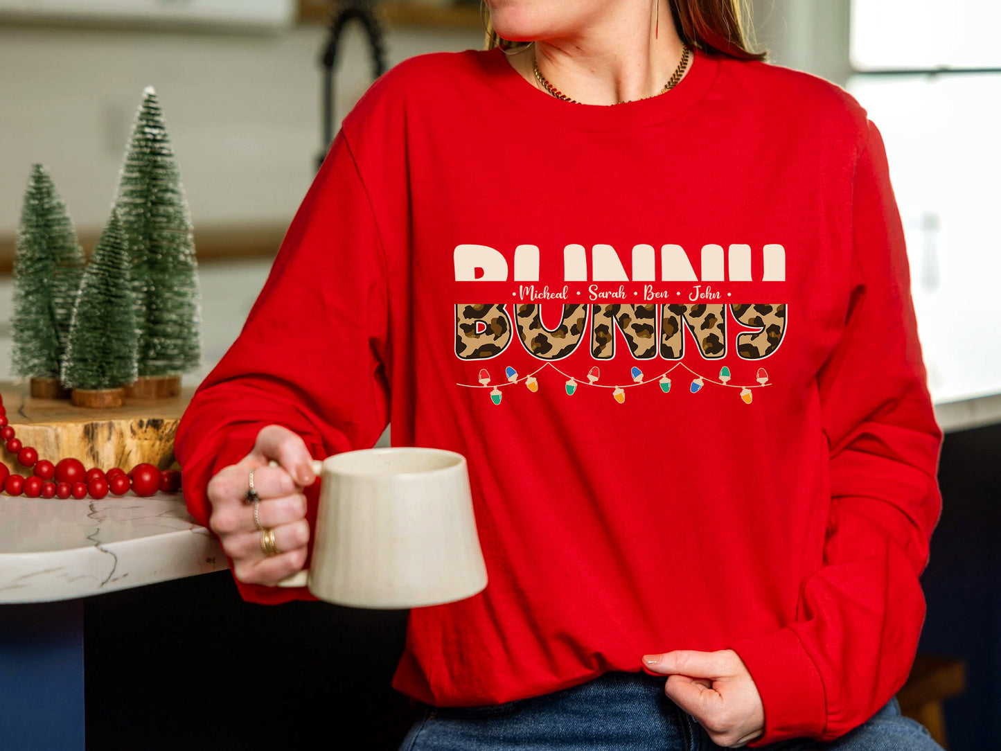 A person wearing a red sweater with the text "BUNNI" printed on it, holding a white mug and standing in a kitchen setting with Christmas decorations visible in the background.