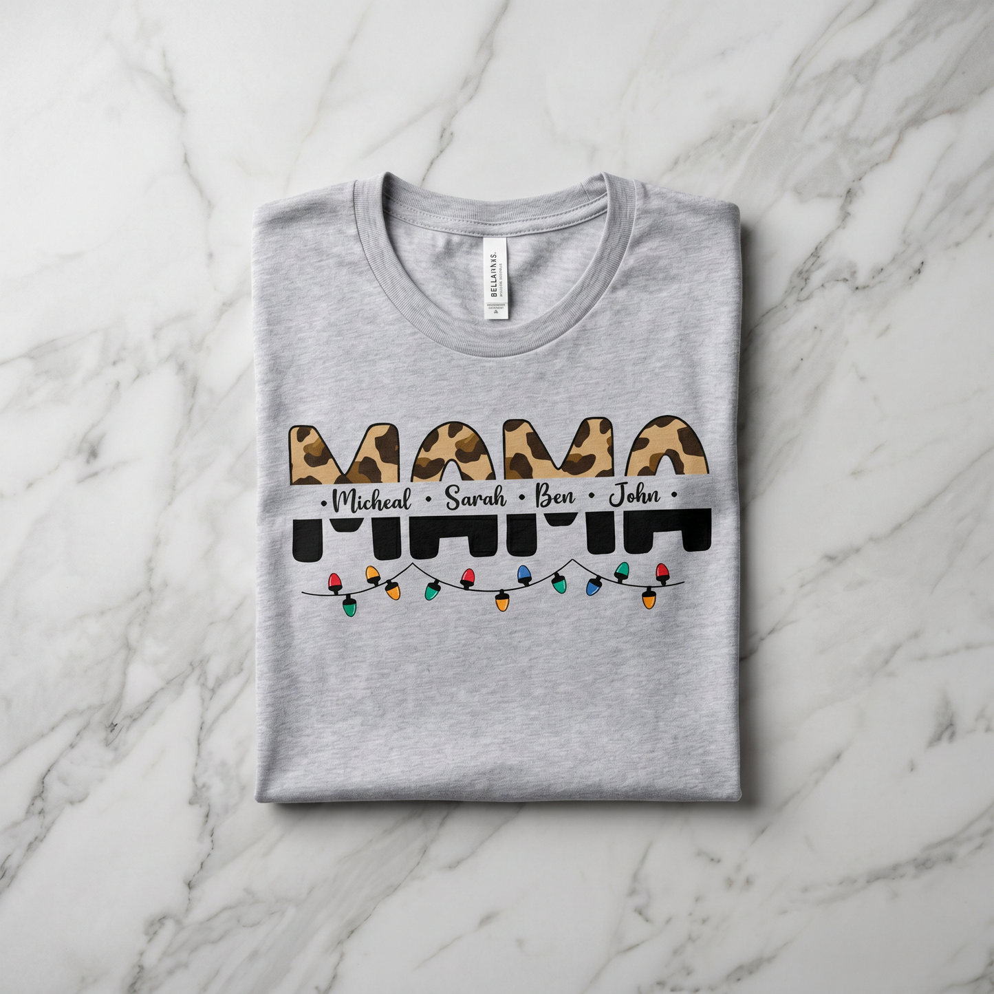 A gray t-shirt with a graphic design featuring the word "MAMA" in a leopard print font, along with the names "Michael", "Sarah", and "Ben" written below it. The design also includes colorful Christmas lights.