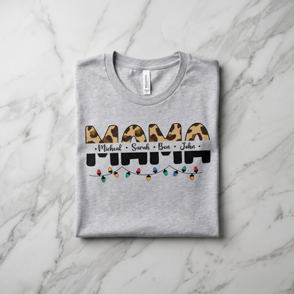 A gray t-shirt with a graphic design featuring the word "MAMA" in a leopard print font, along with the names "Michael", "Sarah", and "Ben" written below it. The design also includes colorful Christmas lights.