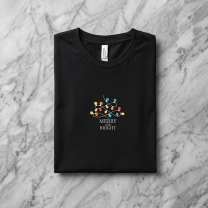 A black t-shirt with a graphic design on it, featuring a Christmas tree with colorful lights and the text "MERRY on BRIGHT" printed on it.