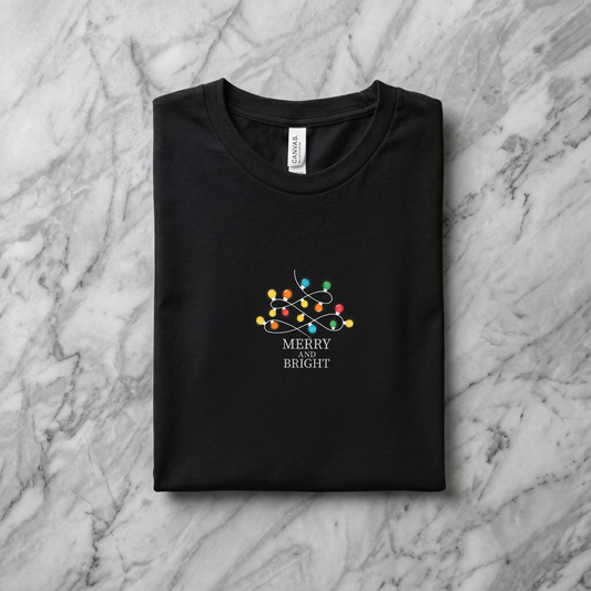 A black t-shirt with a graphic design on it, featuring a Christmas tree with colorful lights and the text "MERRY on BRIGHT" printed on it.