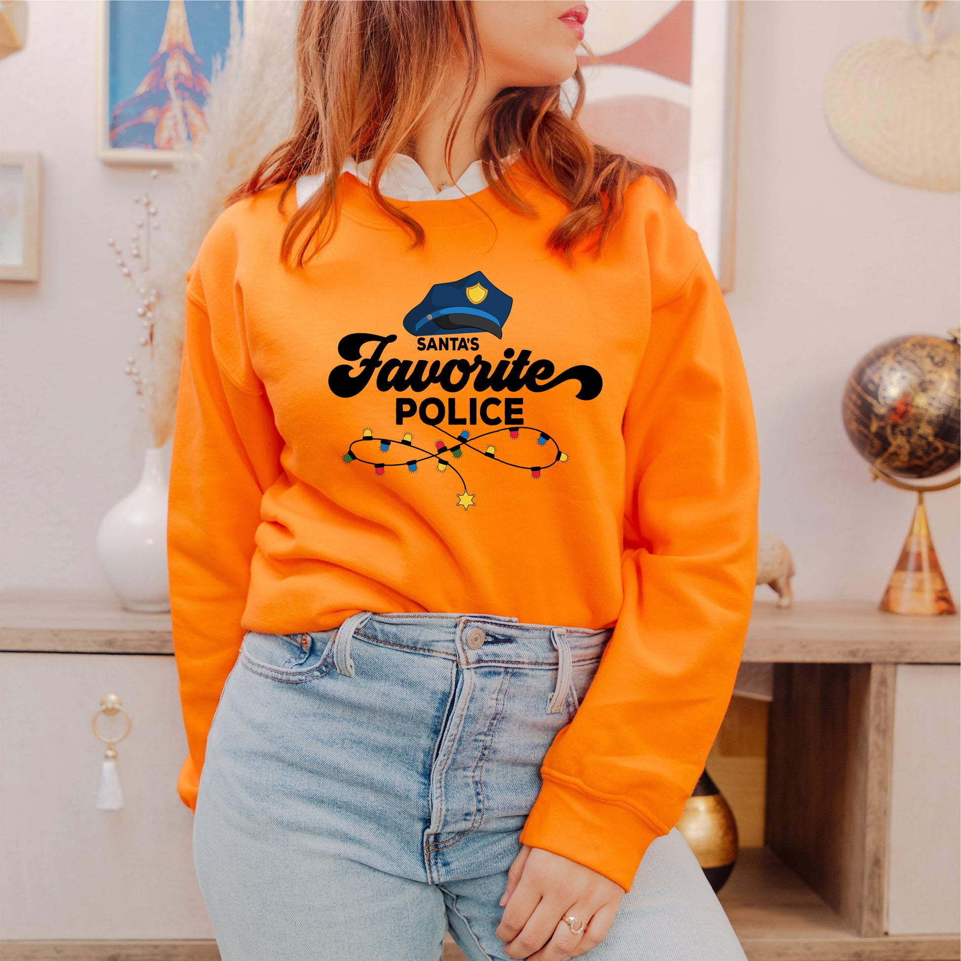 A woman wearing an orange sweatshirt with a graphic of a police officer and the text "Santa's Favorite Police" is standing in a room with various decor items visible in the background.