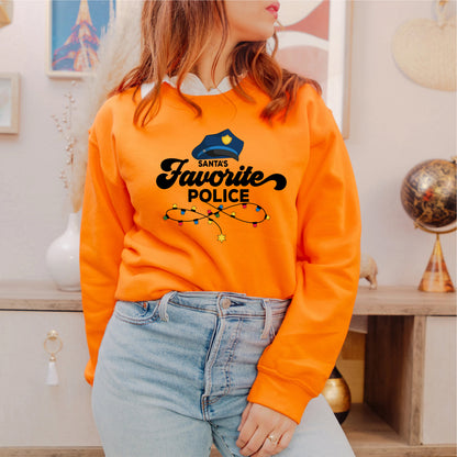 A woman wearing an orange sweatshirt with a graphic of a police officer and the text "Santa's Favorite Police" is standing in a room with various decor items visible in the background.
