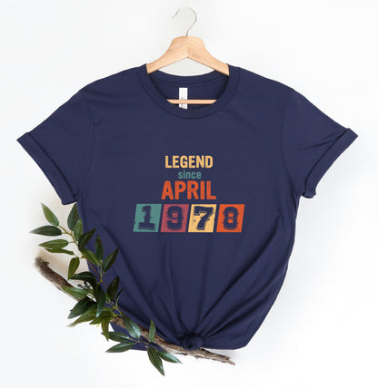 A navy blue t-shirt with the text "LEGEND since APRIL 1978" printed on it, along with a colorful block design. The t-shirt is hanging on a wooden hanger, and there is a branch with green leaves next to it.