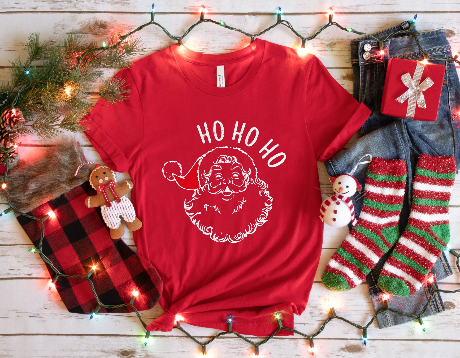 A red t-shirt with a Santa Claus design and the text "HO HO HO" is displayed on a wooden surface, surrounded by Christmas decorations such as a gingerbread man, a gift box, and striped socks.