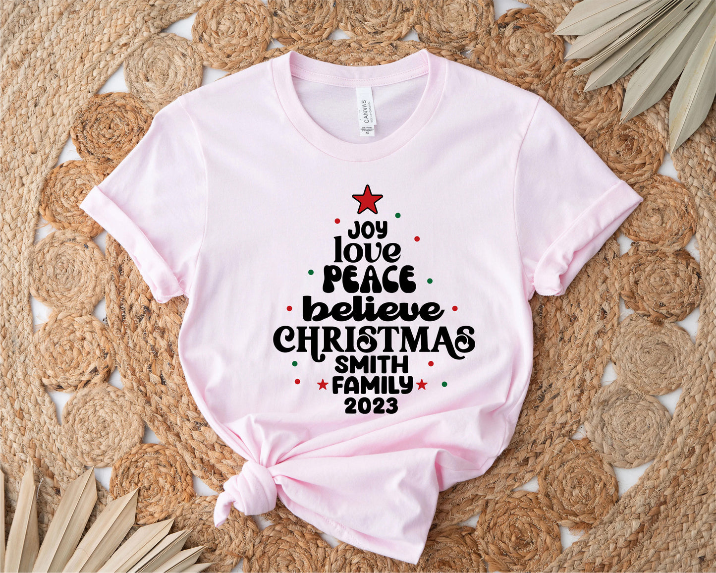 A pink t-shirt with a Christmas-themed design printed on it, placed on a woven mat with palm leaves.