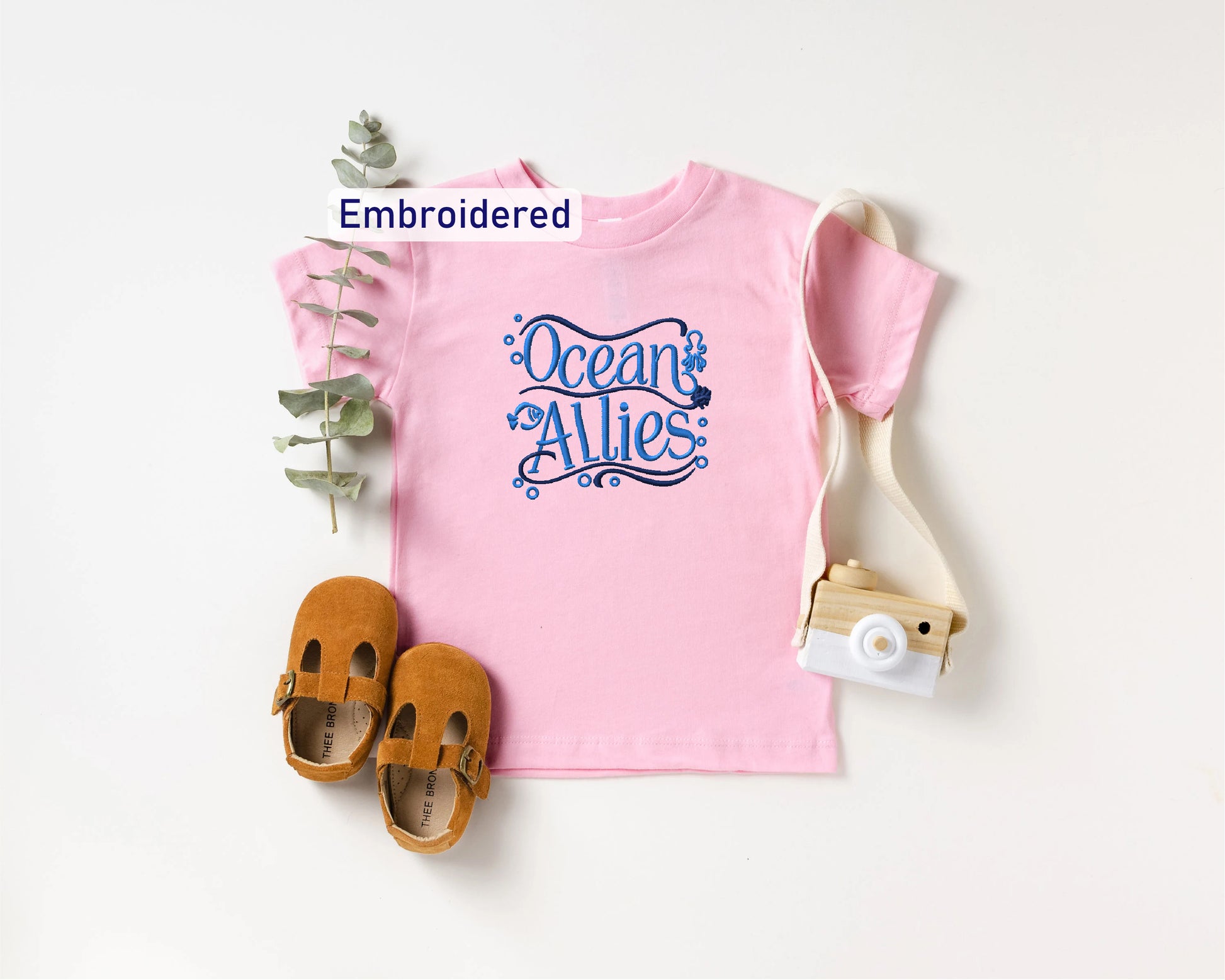 a pink t-shirt with a blue design, a pair of brown baby shoes, and a white camera, all arranged on a white background.