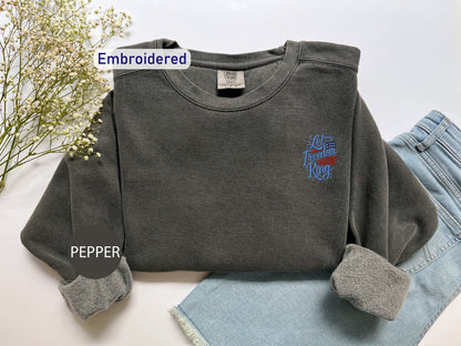 a gray sweatshirt with an embroidered design on the left chest area, placed on top of a pair of blue jeans.