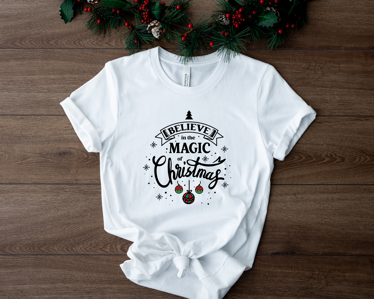 A white t-shirt with a printed design on it, featuring the text "Believe in the Magic Christmas" surrounded by Christmas-themed elements such as ornaments and pine branches.