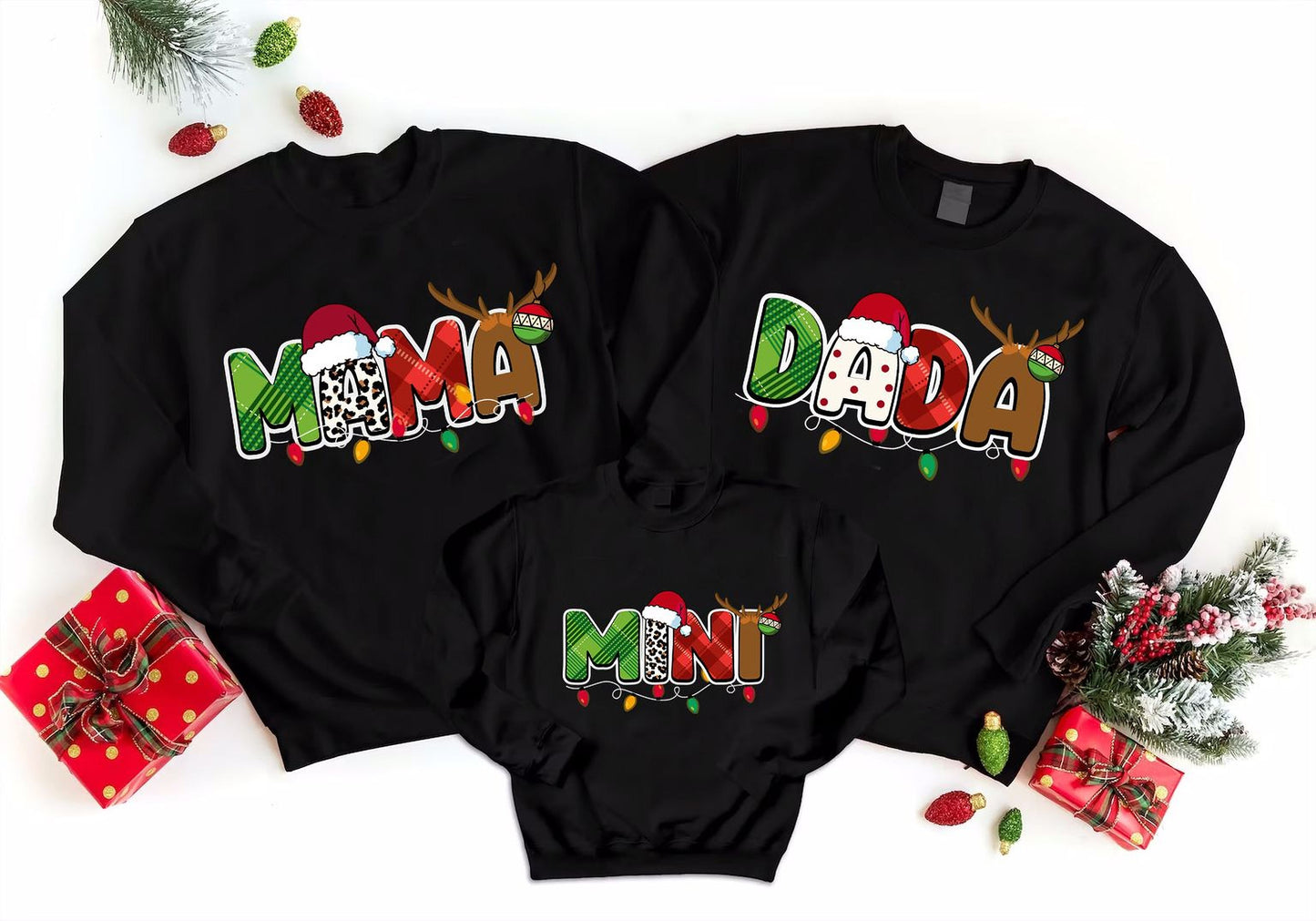 A collection of three black sweatshirts with the word "MAMA" printed on them, along with the names "DADA" and "MINT" printed below. The sweatshirts are arranged on a white background, and there are Christmas decorations, including ornaments and a small Christmas tree, surrounding them.