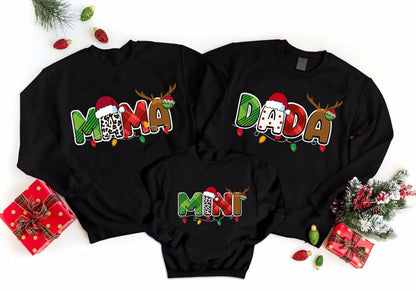 A collection of three black sweatshirts with the word "MAMA" printed on them, along with the names "DADA" and "MINT" printed below. The sweatshirts are arranged on a white background, and there are Christmas decorations, including ornaments and a small Christmas tree, surrounding them.