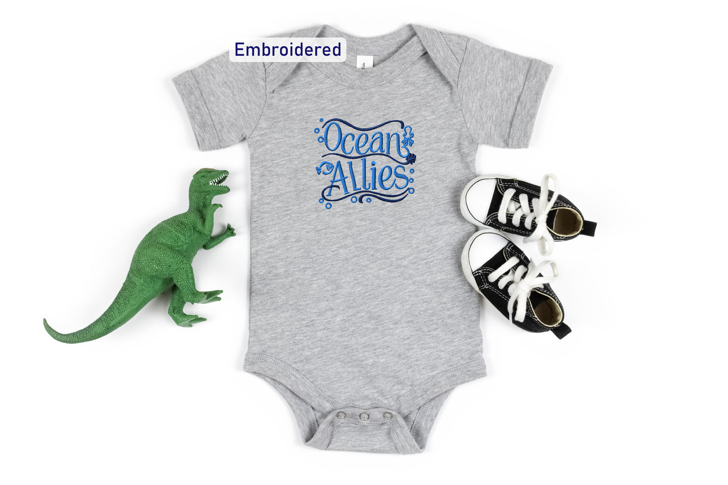 a gray onesie with the text "Ocean Allies" embroidered on it, placed next to a green dinosaur toy and a pair of black and white sneakers.