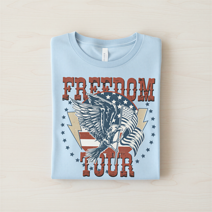 A light blue t-shirt with a graphic design featuring an eagle, an American flag, and the text "FREEDOM TOUR" printed on it.