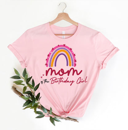 A pink t-shirt with a rainbow graphic and the text "Mom of the Birthday Girl" printed on it, along with a plant branch and leaves.