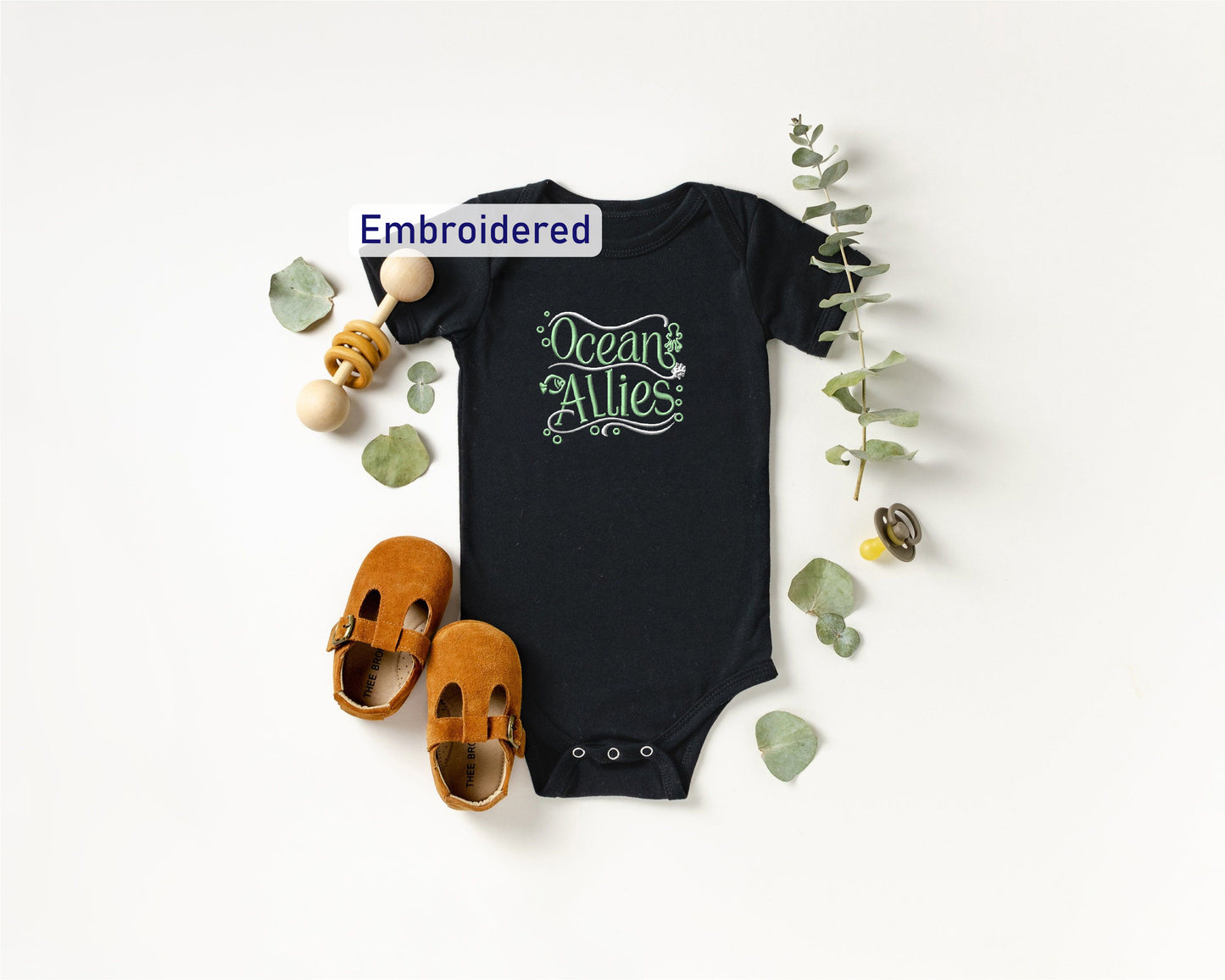 a black baby onesie with the text "Ocean Allies" embroidered on it, along with a pair of brown baby shoes and some green leaves.