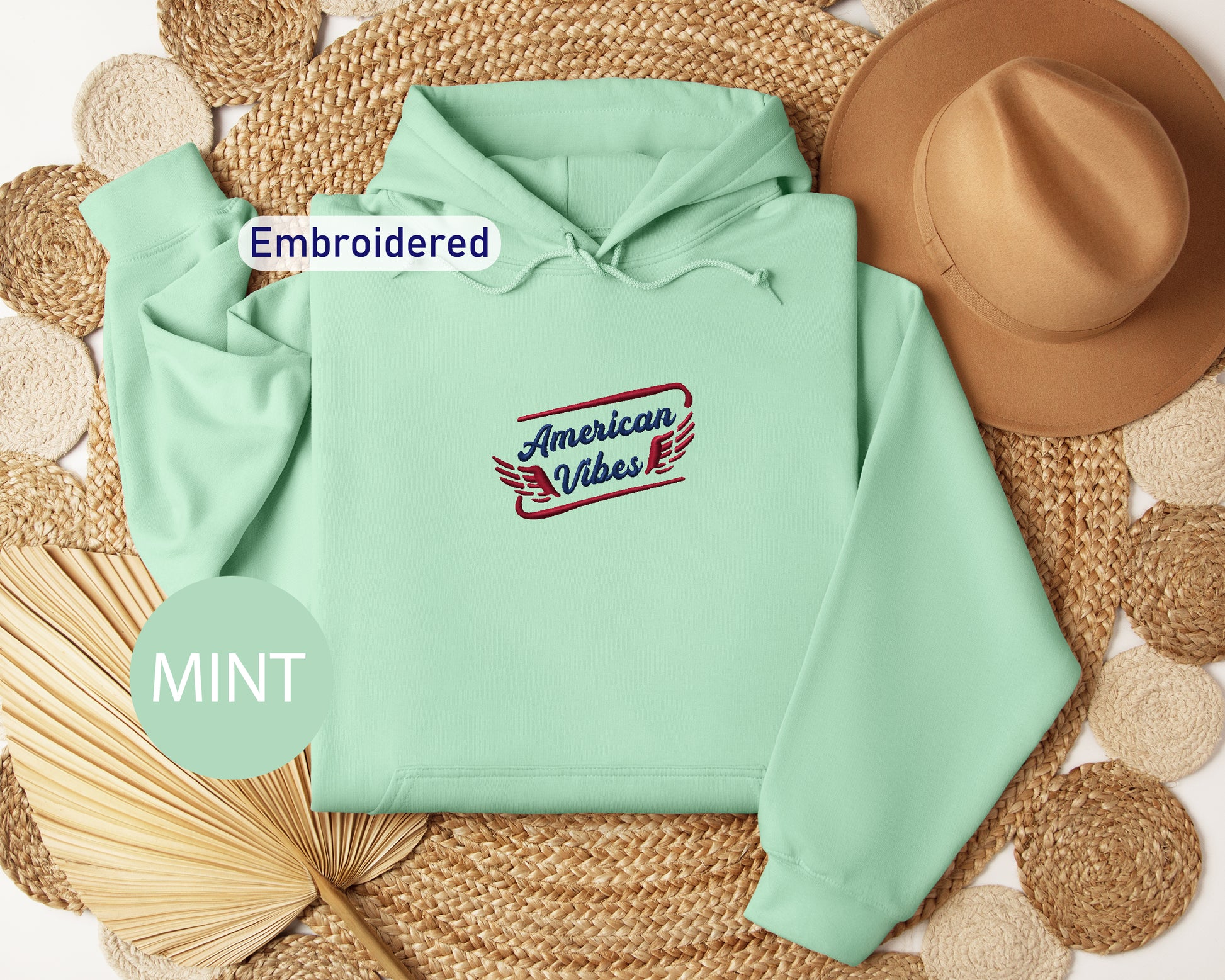 a mint green hoodie with an embroidered design, a brown hat, and a straw hat, all arranged on a woven mat.
