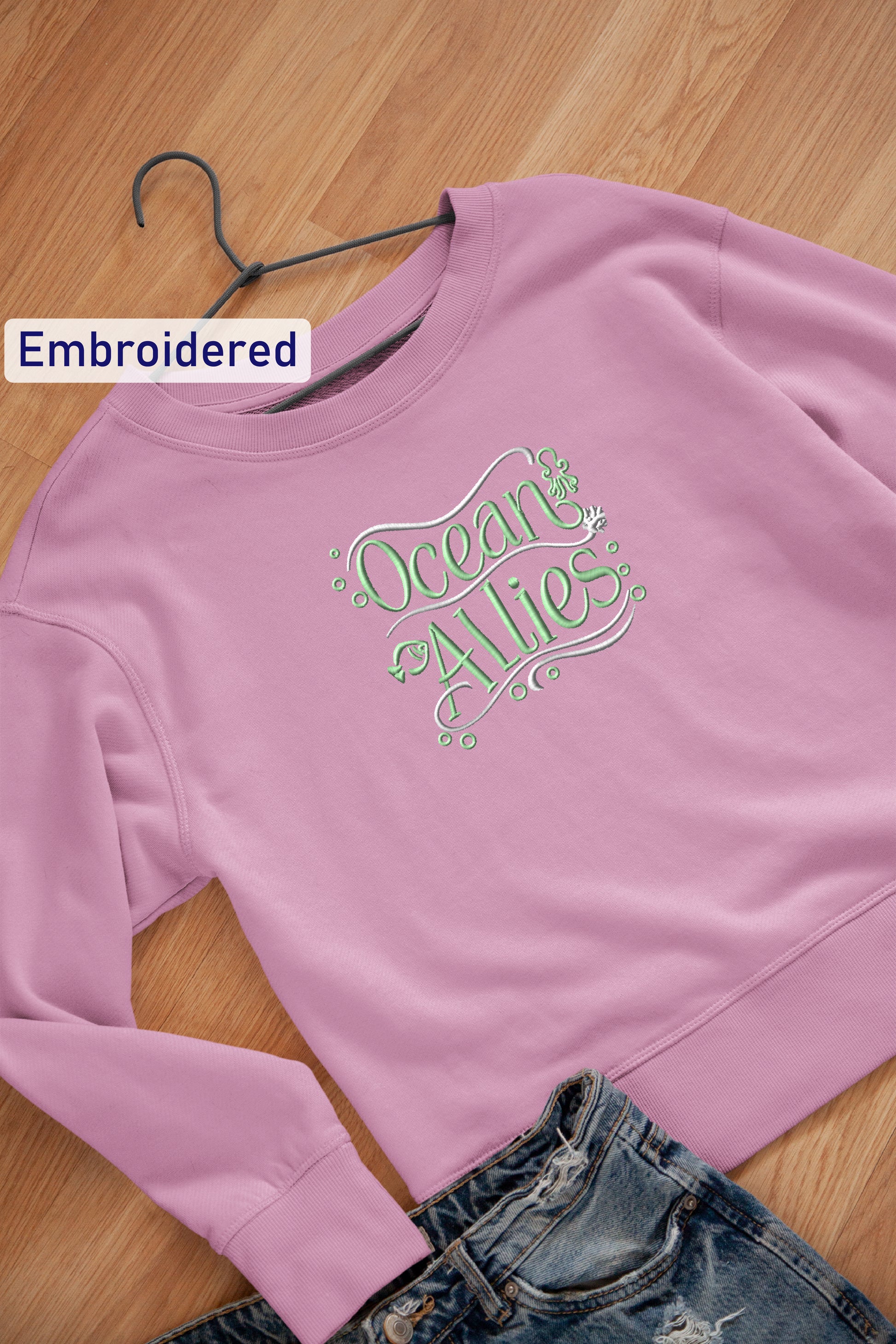 A pink sweatshirt with the text "Ocean Allies" embroidered on it, along with a pair of blue jeans, are laid out on a wooden surface.