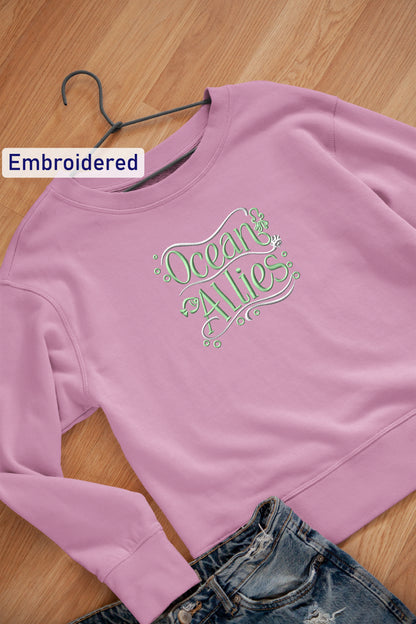 A pink sweatshirt with the text "Ocean Allies" embroidered on it, along with a pair of blue jeans, are laid out on a wooden surface.