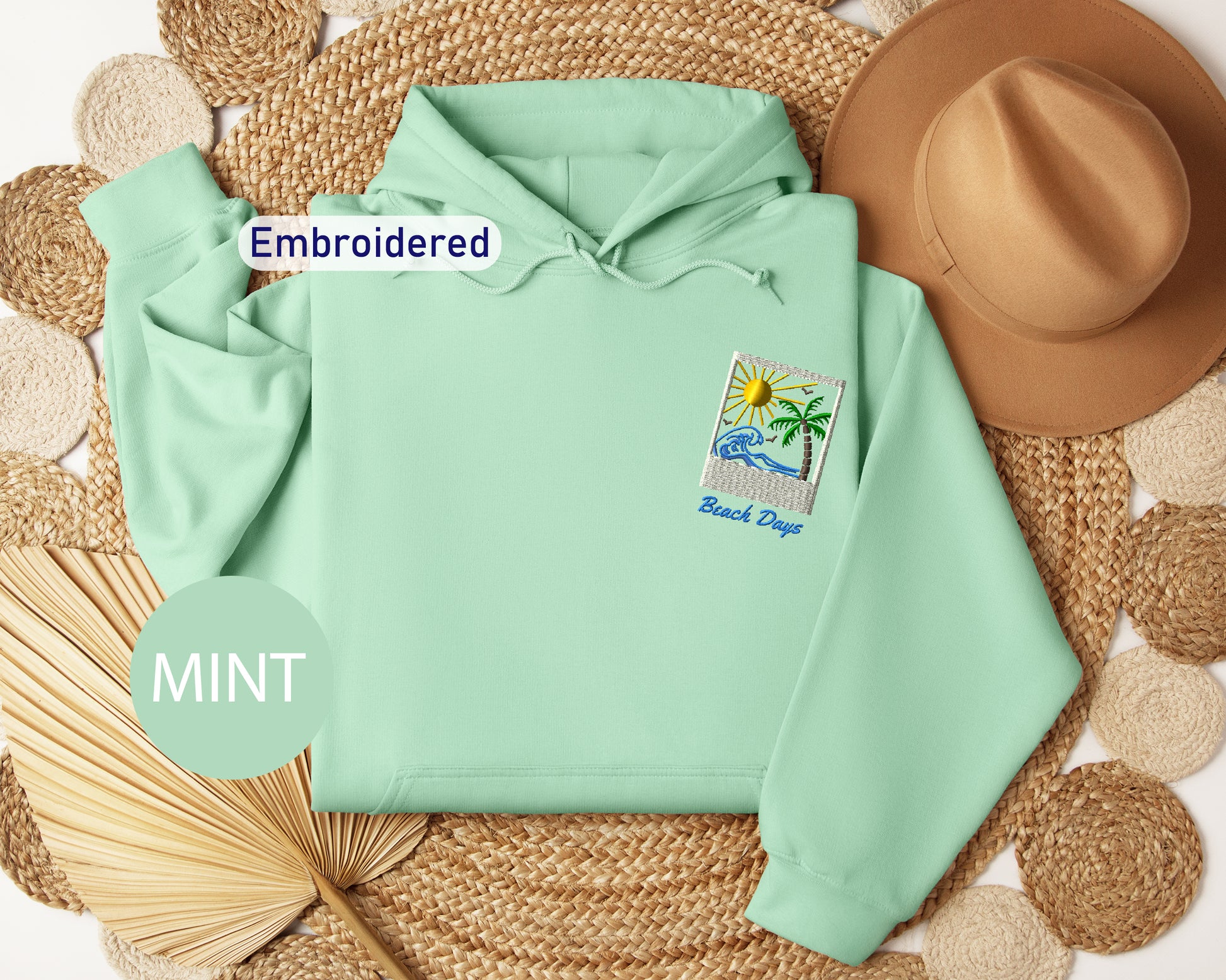 a mint green hoodie with a beach-themed patch on the left chest, a brown hat, a straw hat, and a woven mat on the right side.