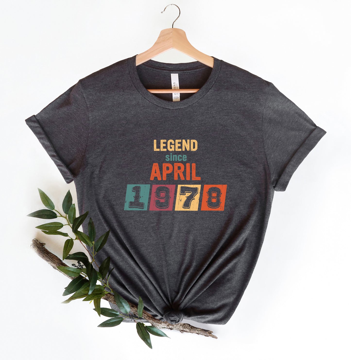 A gray t-shirt with the text "LEGEND since APRIL 1978" printed on it, along with a colorful graphic design. The t-shirt is hanging on a wooden hanger, and there is a branch with green leaves next to it.