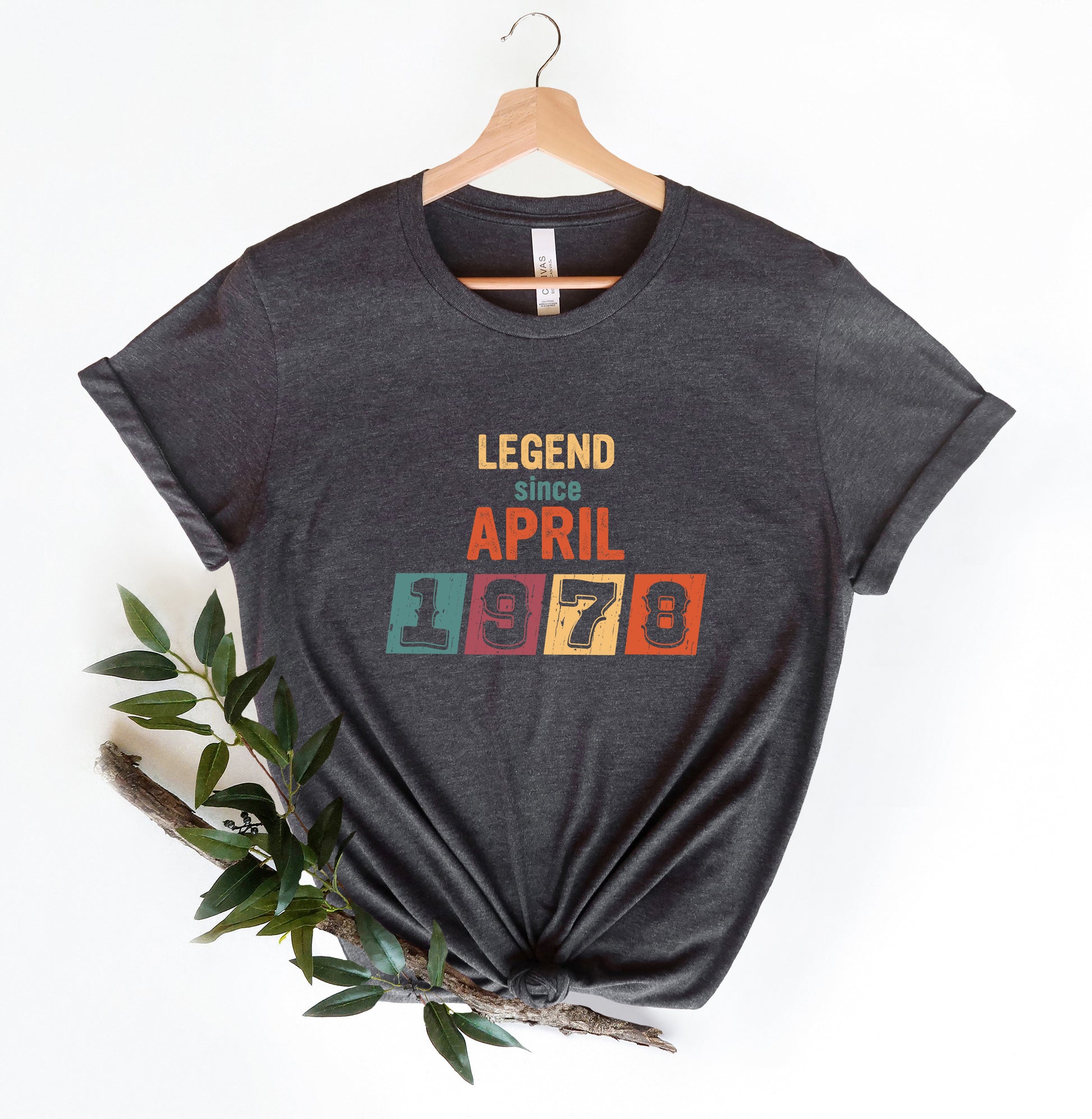 A gray t-shirt with the text "LEGEND since APRIL 1978" printed on it, along with a colorful graphic design. The t-shirt is hanging on a wooden hanger, and there is a branch with green leaves next to it.