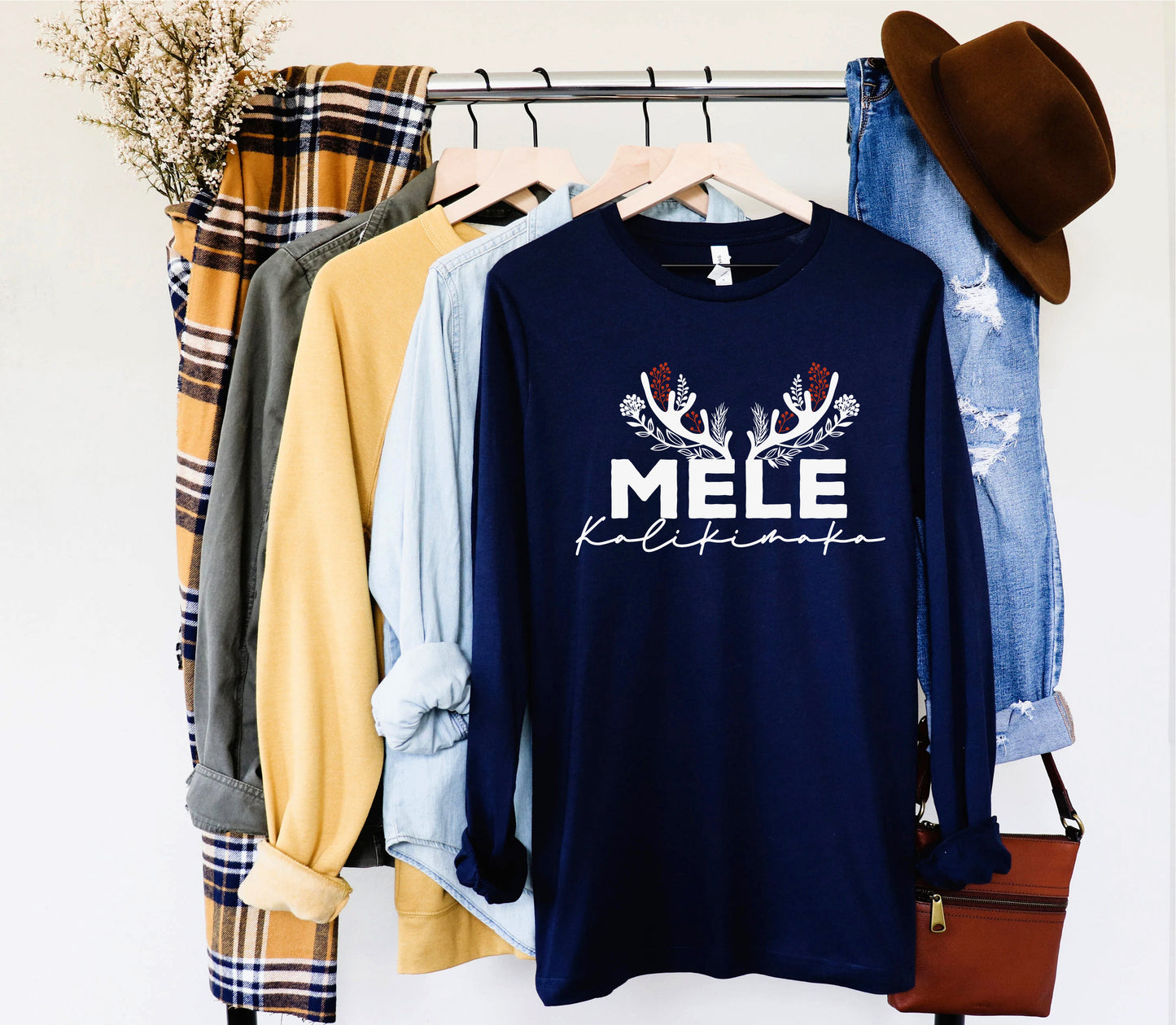A clothing rack displaying various items, including a navy blue long-sleeve shirt with the text "MELE KALIKINMAK" printed on it, a plaid shirt, a yellow shirt, and a pair of blue jeans.